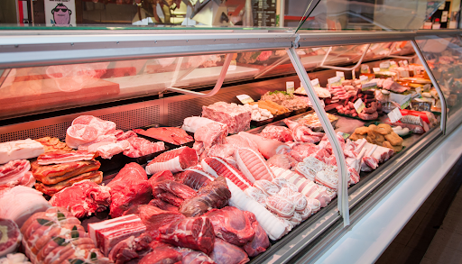 A wide-angle shot of a refrigerated butcher's counter filled with various cuts of raw red meat, sausages, and other meat products.